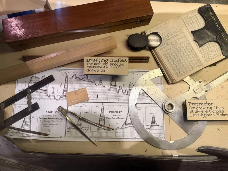 The full surveying case: wooden instrument box, drafting scales, a magnifying loupe, the leather-bound Engineer's Pocket Table-Book propped open, and the big protractor with its hand-lettered card — "for drawing lines at different angles (120 degrees shown)."