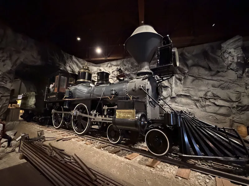 The whole engine in its rock-walled diorama: black boiler, balloon stack, giant oil headlamp, cowcatcher raking forward, gold "Gov. Stanford" lettering on the cab, and a mannequin engineer leaning from the window. A 4-4-0 American type, the classic silhouette of the era.