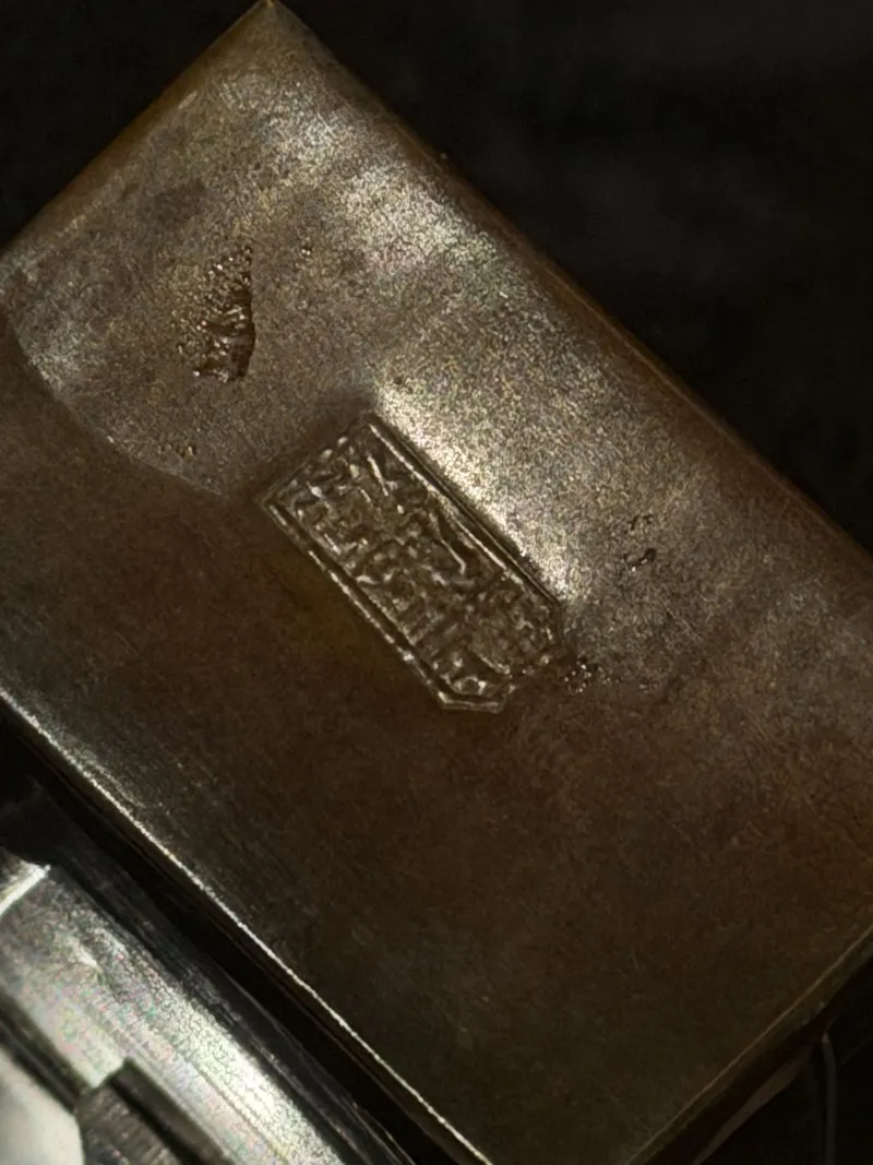 A macro of a brass opium box — the kind archaeologists find by the hundreds at Central Pacific work camps, among the most common artifacts of the Chinese railroad workers' daily lives. The rectangular cartouche is a Chinese maker's or brand stamp, characters running in vertical columns. We worked the image hard — rotation, relief filtering, glare removal — but between a century of wear and the case lighting's glare, the individual characters can't be honestly read from this photo; a reading would be a guess. Stamps like this typically carry the maker's shop or brand name — the licensed Hong Kong brands Fook Lung and Lai Yuen marked their containers exactly this way. A raking-light photo on a return visit would likely crack it.