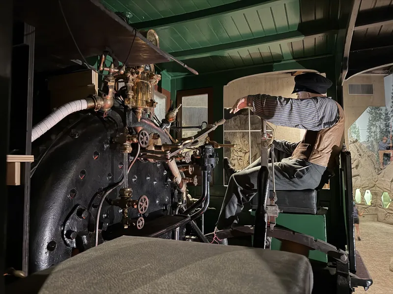 Inside the Gov. Stanford's cab: green-painted woodwork, the black riveted backhead crowded with brass — pressure gauge on its curved pipe, tall ornate lubricator, wheeled valves — the long Johnson bar with its notched quadrant, and a mannequin engineer at his armrest watching the road.