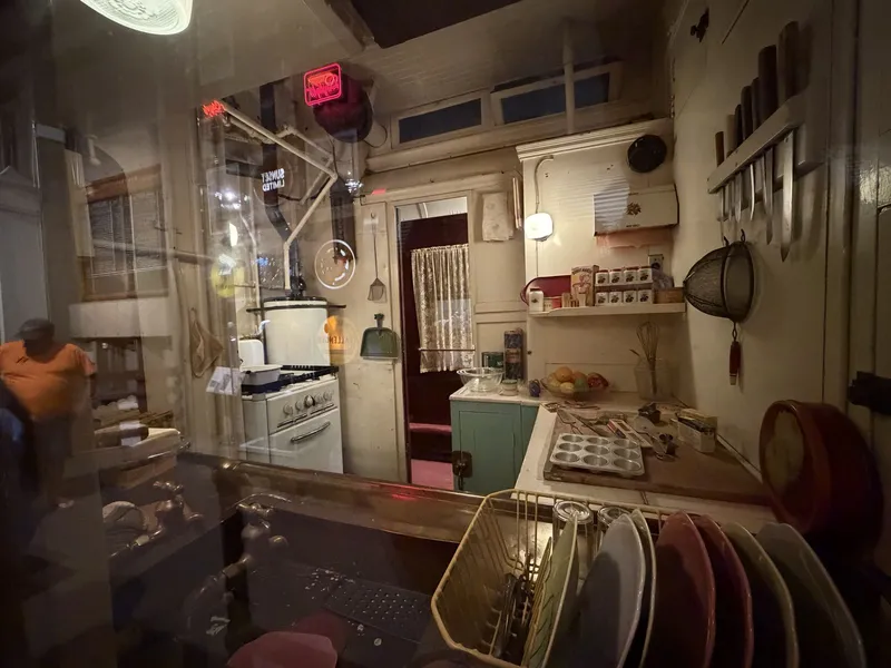 A dining-car galley preserved mid-shift: white enamel stove, muffin tin on the counter, knives on the wall rack, pastel plates in the drying rack, and the Sunset Limited's window lettering reading backwards through the glass, neon glowing beyond.