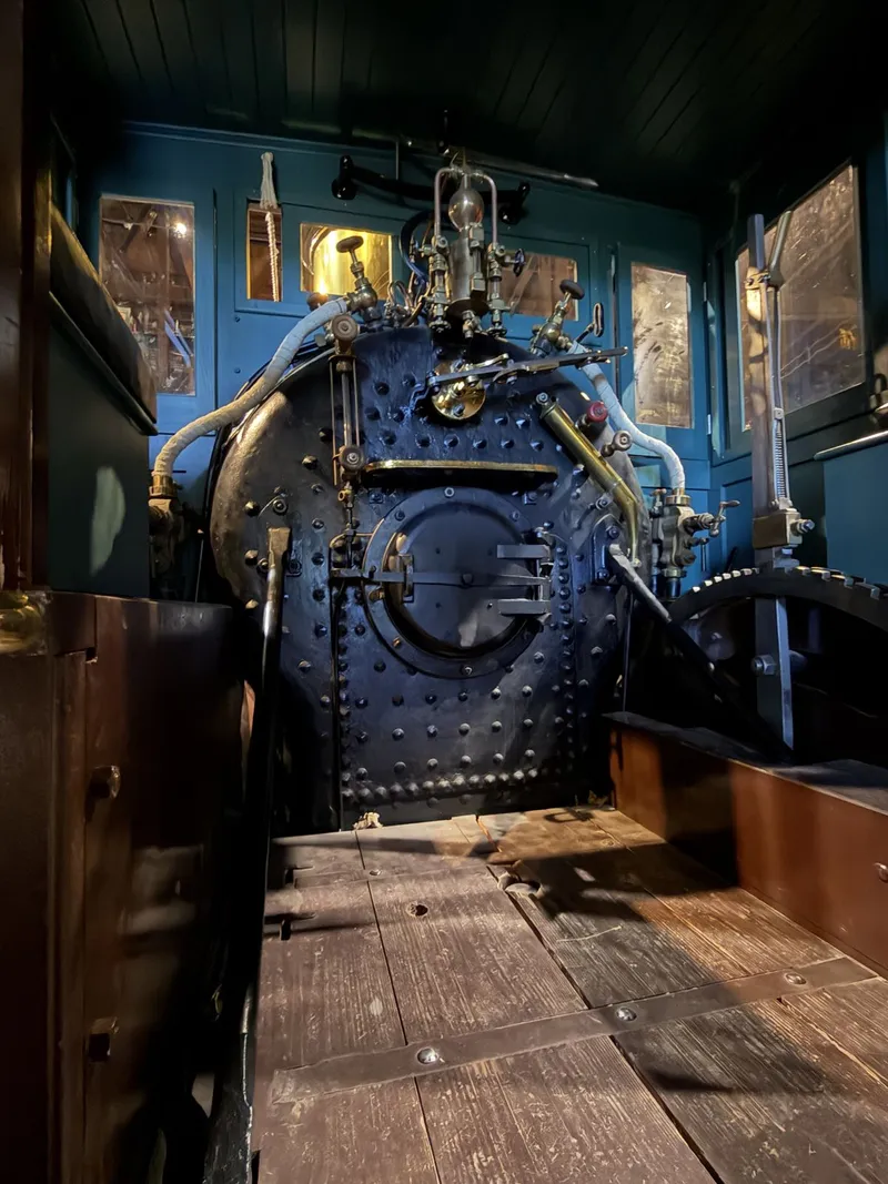 Inside a wood-burner's cab, painted teal: the black riveted backhead like a studded door, the furnace door at its center, white-lagged steam pipes snaking to brass fittings, and daylight through the little arched windows.