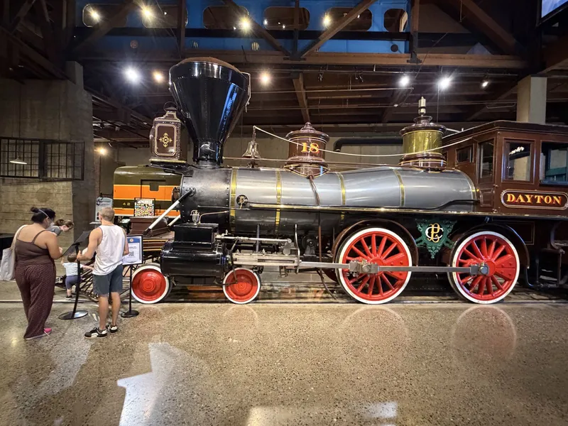 The Dayton, museum visitors at the rope for scale: silver boiler, bright red wheels, the green-and-gold CP monogram on her frame, "DAYTON" in gold on the cab panel. An engine built to be admired as much as worked.