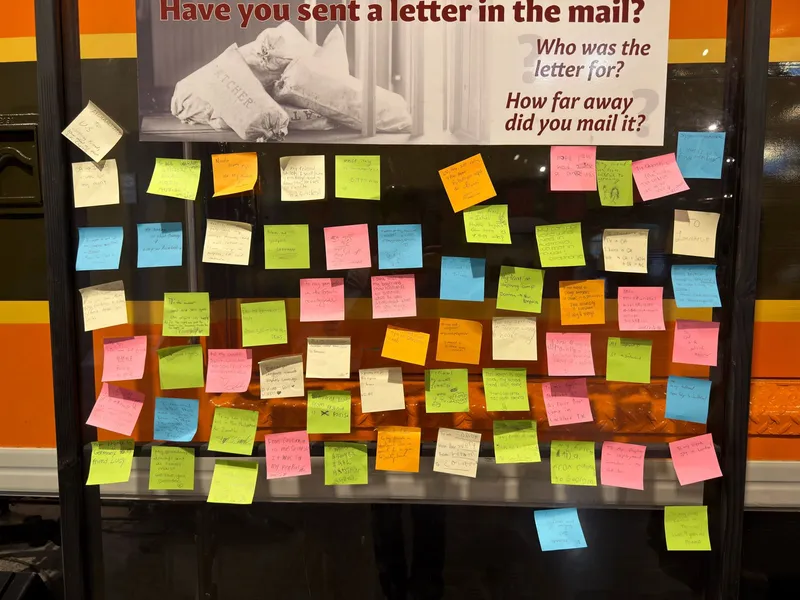 The visitor wall: "Have you sent a letter in the mail? Who was it for? How far away?" — answered in dozens of sticky notes. Afghanistan to the U.S. Fiji. Venice to Sacramento, 5,999 miles. A boyfriend stationed in Bahrain, now a husband. A daughter at Air Force boot camp. A grandma who always sent "snail mail."