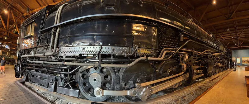 The panorama: 4294's number on the cab — at the front, which is the whole point — then a city block of black boiler, silver side rods, and disc drivers with little SP stars, curving away down the hall in the wide lens.