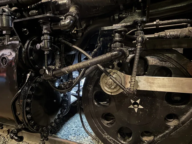 Close in on the running gear: the polished side rod meeting a crank pin with its big hex nut, a white star on the black disc driver, and the famous tangle of wrapped and armored lines — most of them, it turns out, carrying oil.