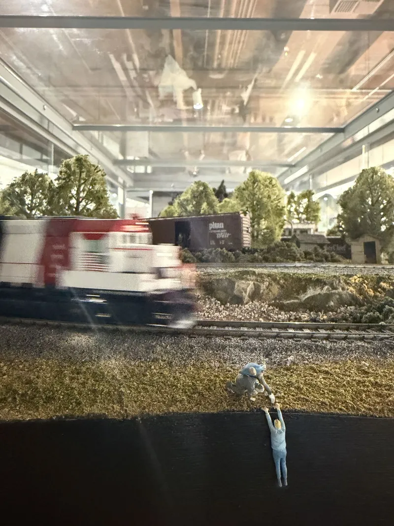 The layout's best joke: a red-and-white diesel blurring past, and in the foreground two figures at the very edge of the benchwork — one dangling off the fascia into the void, his buddy hauling him back up. Model builders having fun with the fourth wall.