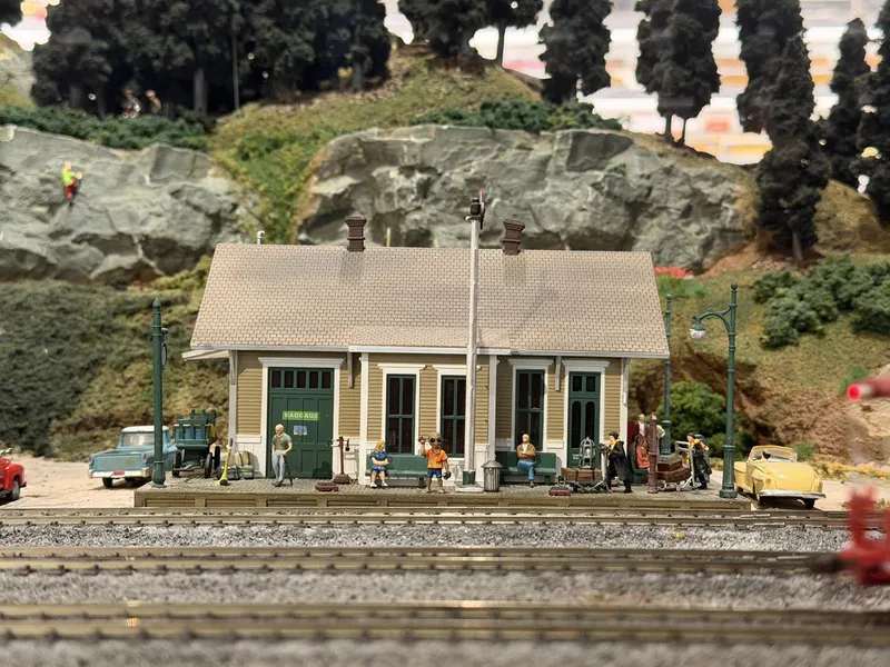 The model railroad's little depot: tan station with a green BAGGAGE door, figures waiting on the platform — including one gentleman in a loud Hawaiian shirt — a blue pickup and a yellow convertible parked outside, and if you look at the painted rocks above, a tiny climber halfway up the cliff.