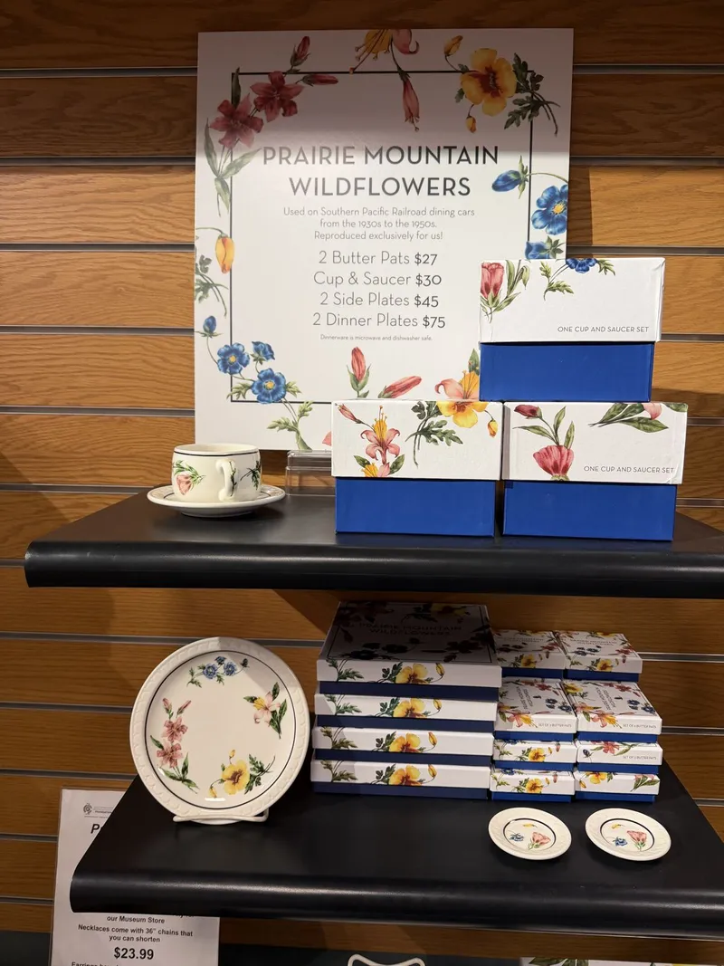 The gift shop's Prairie Mountain Wildflowers display: the reproduction china Southern Pacific ran in its dining cars from the 1930s to the 1950s — a cup and saucer on the shelf, floral-printed boxes stacked below, butter pats at $27 the pair.