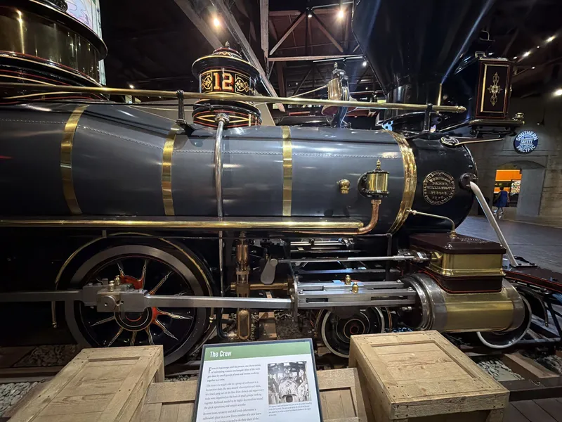 No. 12 in profile: slate-gray boiler with polished brass bands, the ornate sand dome painted with a gold "12," red-and-gold pinstriped wheels, brass-jacketed cylinder, and the Baldwin plate on her smokebox. Victorian machine-as-jewelry.